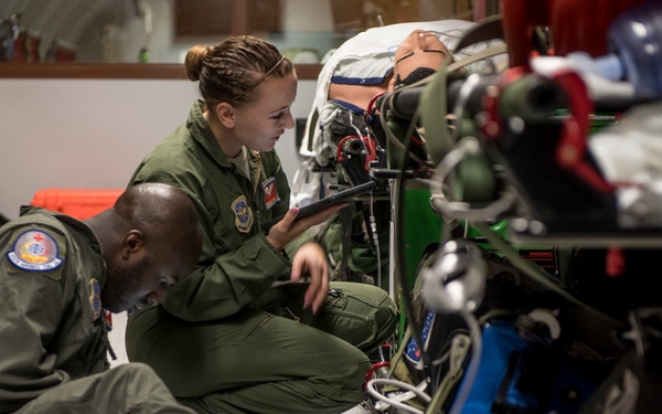 Two military personnel in olive green flight suits are working together on medical equipment, likely inside a military aircraft or training facility. They appear to be inspecting or configuring a complex aeromedical evacuation litter system with multiple medical devices attached. A simulated patient mannequin is visible on the litter, suggesting this may be a training exercise.