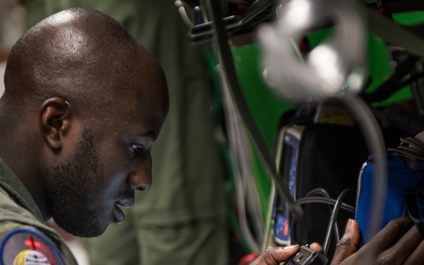 A military airman in an olive green flight suit is focused on operating a tablet device in what appears to be an aircraft or equipment bay. He is wearing a unit patch on his left shoulder, suggesting he is an active duty service member performing a technical or operational task. The environment contains multiple cables, connectors, and electronic equipment consistent with an aviation or maintenance setting.