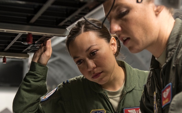 Two female U.S. Air Force personnel in olive green flight suits are reviewing documents together in what appears to be the interior of a military aircraft. The individual on the left, whose name tag reads 'CRYSTAL L...', is wearing a patch indicating Air Mobility Command. Both appear focused on the paperwork being reviewed.