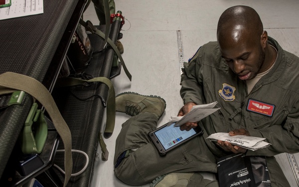A military personnel member wearing an olive green flight suit with US flag patch and wing insignia is seated on the floor reviewing documents and a tablet device. They are surrounded by various medical and tactical equipment including a blood pressure cuff, monitoring devices, and supply cases. The setting appears to be an indoor facility, likely during equipment inspection or mission preparation.