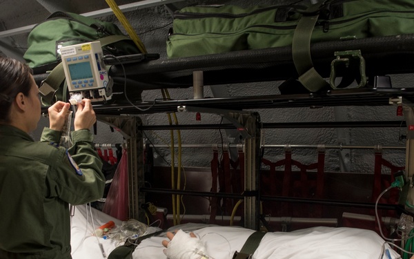 A person wearing an olive green military flight suit is operating medical monitoring equipment mounted above a litter (military stretcher) in what appears to be a military aeromedical evacuation training environment. A medical training mannequin is strapped to the litter, connected to various tubes, IV lines, and a cervical collar, simulating a patient in transport. The setting appears to be a ground-based aeromedical evacuation training simulator or mock aircraft interior.