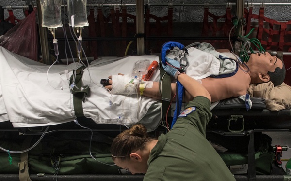 A U.S. military medical personnel in olive green flight suit is performing aeromedical evacuation training inside what appears to be a military transport aircraft. She is attending to a medical training mannequin on a litter/stretcher equipped with IV lines, monitoring equipment, and a breathing mask. Various medical devices including a portable monitor/defibrillator are visible on the floor.