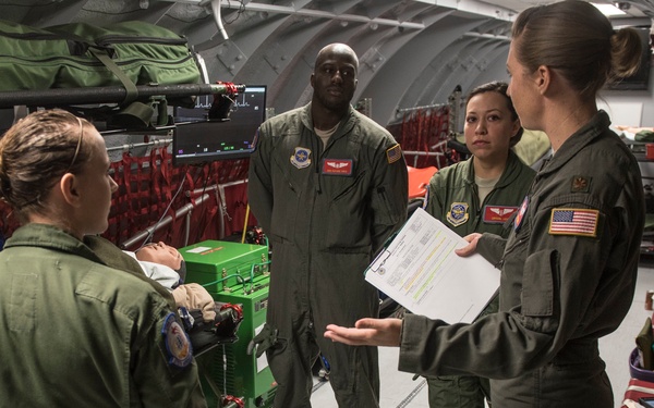 Four military personnel in olive green flight suits are gathered inside what appears to be a military aeromedical evacuation aircraft. One person is holding a clipboard with documents while the group appears to be conducting a briefing or patient handoff discussion. Medical equipment, litters, and supplies are visible throughout the aircraft interior, along with what appears to be a patient on a litter in the background.