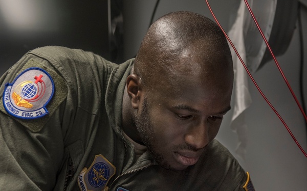 A U.S. Air Force airman identified as SrA Kayode Idris from the 315th Aeromedical Evacuation Squadron is performing a medical procedure on a training mannequin arm. He is wearing an olive green flight suit with unit patches and purple nitrile gloves, practicing what appears to be an intravenous (IV) insertion technique. The setting appears to be inside a military aircraft or simulation environment.