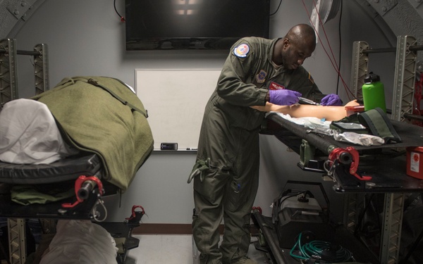 A military medical personnel wearing an olive green flight suit and purple nitrile gloves is performing a medical procedure or training exercise on what appears to be a medical training mannequin arm on a litter/stretcher rack. The setting appears to be the interior of a military aircraft or a simulator replicating one, equipped with multiple litter stanchion systems. A patient or mannequin covered with a white sheet and olive blanket is visible on a litter to the left.