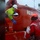The image shows a maritime rescue or boarding operation involving what appears to be US Coast Guard personnel in red survival suits and helmets interacting with a crew member in a yellow high-visibility jacket and white hard hat on a red vessel. The vessel has partial text visible reading 'RESOL...' and 'DE' on its hull. Conditions appear rough and wet, suggesting an active sea rescue or inspection operation.