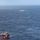 An aerial photograph shows two vessels operating in open ocean under moderately choppy conditions. In the foreground is a red salvage/rescue tug bearing the name 'RESOLVE,' while in the background is a larger white vessel consistent with a Coast Guard cutter. The two ships appear to be operating in proximity, suggesting a coordinated maritime operation.