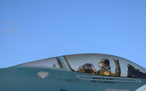 A military jet fighter, numbered 335, is shown on the ground with a pilot seated in the cockpit giving a hand gesture. The aircraft bears the names 'ANDREW HONG' and 'ROB VILLARREAL' stenciled below the canopy, indicating assigned crew members. The aircraft appears to be an F-16 Fighting Falcon in a teal/blue-gray paint scheme, consistent with aggressor squadron livery.