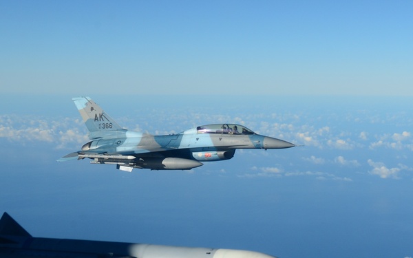 An aerial photograph showing a U.S. Air Force F-16 Fighting Falcon jet fighter in flight, marked with 'AK' tail code and serial number 366, indicating it belongs to an Alaska-based unit. The image is taken from another aircraft, with a missile or external store visible in the foreground. The aircraft is flying over what appears to be a mountainous coastal region, likely Hawaii, with scattered clouds and ocean visible in the background.