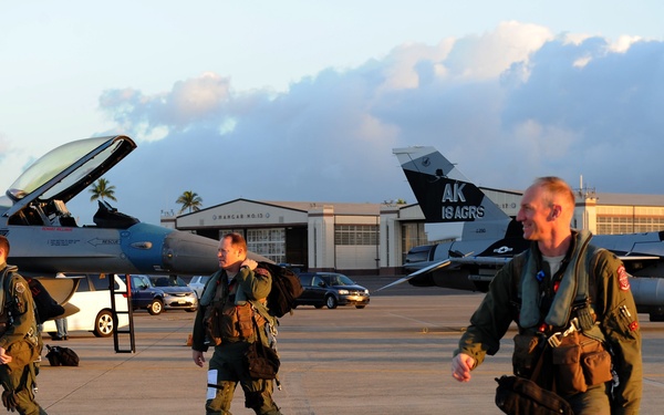 Three military pilots in flight suits and survival gear walk across an airfield tarmac, likely after completing a mission. Behind them are at least two F-16 Fighting Falcon aircraft, one with its canopy open, marked with 'AK' and '18 AGRS' tail codes indicating the 18th Aggressor Squadron based in Alaska. The scene appears to be at a tropical or subtropical air base, likely Hickam Air Force Base in Hawaii, given the palm trees and warm golden lighting.