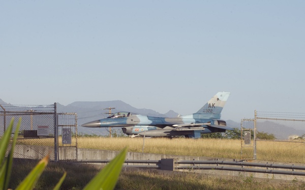 A U.S. Air Force F-16 Fighting Falcon fighter jet with tail code 'AK 322' is visible on or near a taxiway behind a chain-link security fence. The aircraft displays an Alaskan Air National Guard livery in a light blue camouflage scheme. The photo is taken from outside the perimeter fence with tropical vegetation in the foreground and mountains visible in the background.