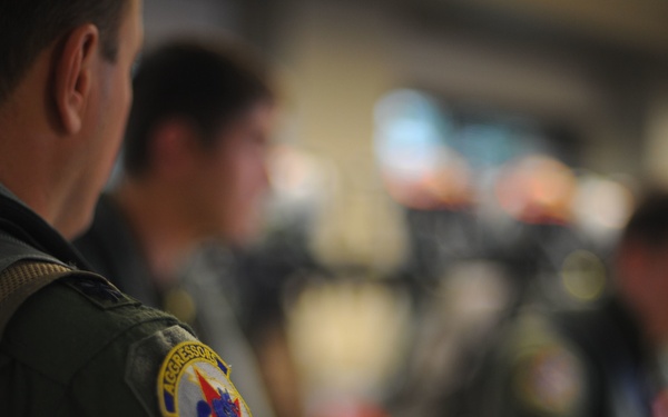 The image shows military personnel in olive/sage green flight suits gathered in what appears to be a briefing room or operations area. The foreground subject displays a clearly visible squadron patch on their left shoulder reading 'AGGRESSORS' with additional text partially visible. Multiple personnel are present in the background, though they are out of focus.