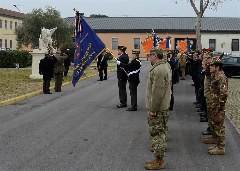 A formal military ceremony is taking place on a paved parade ground, featuring personnel from multiple nations including US and Italian military forces. A color guard is presenting flags, including a prominently visible blue Veterans of Foreign Wars (VFW) of the United States banner. Participants are standing at attention or saluting, suggesting a commemorative or ceremonial event at what appears to be a military installation.