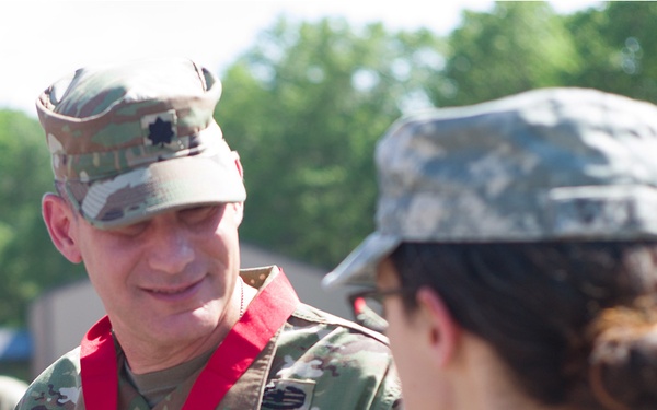 A U.S. Army soldier identified by the name tape 'ADLER' is wearing a red ribbon lanyard with a medal around his neck, suggesting a recent award or recognition ceremony. He is interacting with another soldier in the foreground who appears to be in a different uniform pattern. The setting appears to be an outdoor military environment with trees visible in the background.