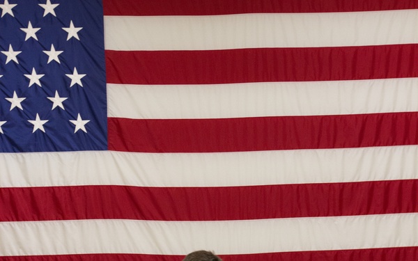 A U.S. Army soldier in OCP (Operational Camouflage Pattern) uniform sits on a metal chair reading documents in front of a large American flag displayed on a wall. The soldier's name tape reads 'PURSWELL' and rank insignia indicates a Captain (O-3). The setting appears to be an indoor military facility, possibly during a ceremony or administrative event.