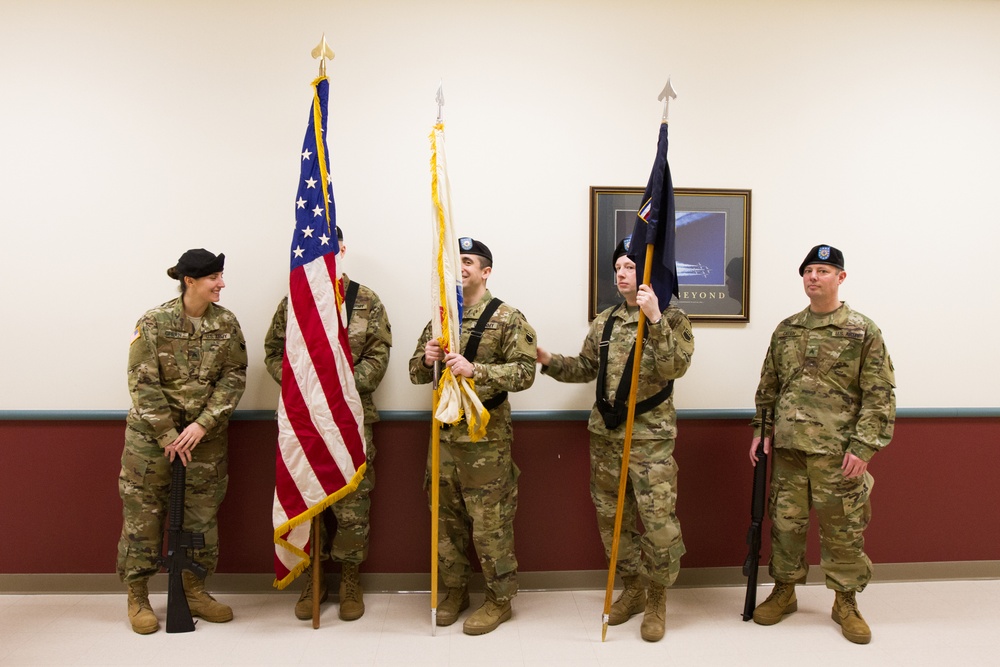 Five U.S. Army soldiers in MultiCam/OCP camouflage uniforms are posed in an indoor military facility, appearing to be a color guard formation. Three soldiers hold ceremonial flag staffs including the U.S. national flag and what appears to be a unit guidon, while two soldiers on the flanks hold rifles at order arms. The group appears relaxed, possibly before or after a formal ceremony.