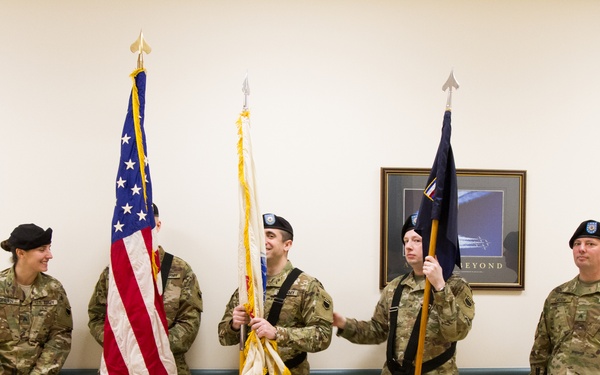 Five U.S. Army soldiers in MultiCam/OCP camouflage uniforms are posed in an indoor military facility, appearing to be a color guard formation. Three soldiers hold ceremonial flag staffs including the U.S. national flag and what appears to be a unit guidon, while two soldiers on the flanks hold rifles at order arms. The group appears relaxed, possibly before or after a formal ceremony.
