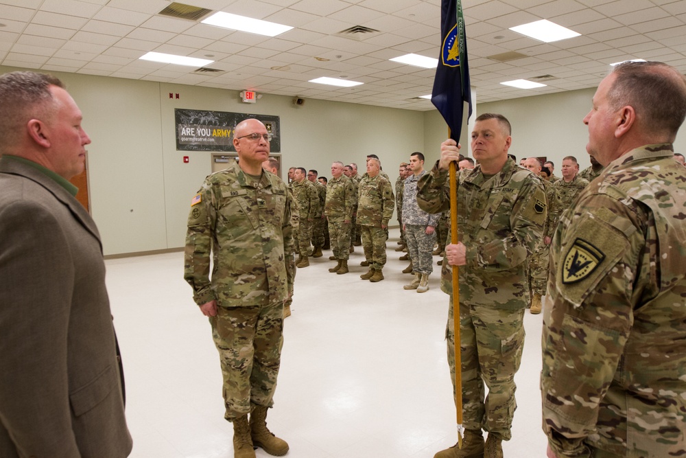 A formal U.S. Army military ceremony is taking place inside an indoor drill hall or armory. Several soldiers in OCP (Operational Camouflage Pattern) uniforms are assembled in formation while a central figure holds a unit guidon/flag. A civilian in a green jacket is also present, suggesting an official change of command or unit activation ceremony.
