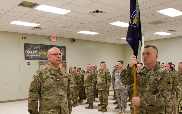 A formal U.S. Army military ceremony is taking place inside an indoor drill hall or armory. Several soldiers in OCP (Operational Camouflage Pattern) uniforms are assembled in formation while a central figure holds a unit guidon/flag. A civilian in a green jacket is also present, suggesting an official change of command or unit activation ceremony.