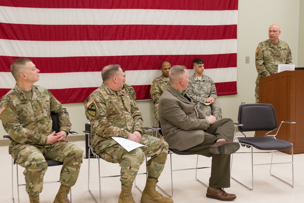 A military ceremony or briefing is taking place indoors with several U.S. Army personnel seated in chairs facing a speaker at a podium. A large American flag is displayed on the wall behind the seated attendees. One civilian in a green jacket is seated among the uniformed military personnel.