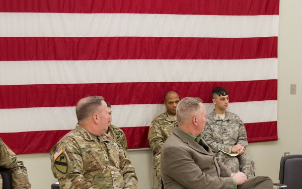 A military ceremony or briefing is taking place indoors with several U.S. Army personnel seated in chairs facing a speaker at a podium. A large American flag is displayed on the wall behind the seated attendees. One civilian in a green jacket is seated among the uniformed military personnel.