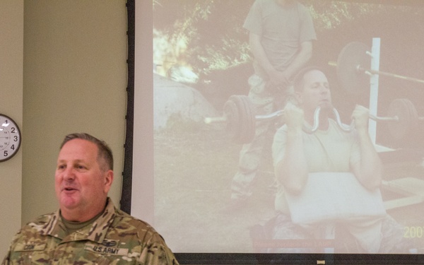 A U.S. Army Colonel is standing at the front of a briefing room, holding papers and apparently giving a speech or presentation. Behind him, a projection screen displays a photograph dated 2007/04/02 showing a person performing a weightlifting exercise outdoors. The setting appears to be a military conference or ceremony room.