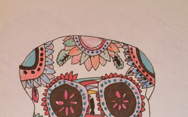 A hand-drawn and colored sugar skull (calavera) artwork created by an artist named Katelyn, as signed in the lower right corner. The skull is decorated in the traditional Día de los Muertos style with intricate floral patterns, geometric designs, and vibrant colors including pink, blue, orange, yellow, and red. The drawing is made on white lined paper using markers or colored pencils.