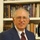An elderly man with gray hair and gold-rimmed glasses poses for a formal portrait wearing a blue blazer, white dress shirt, and red patterned tie. He is seated or standing in front of a well-stocked bookshelf containing numerous religious and theological books. The image appears to be a professional or semi-professional headshot, likely for a church, academic, or organizational profile.