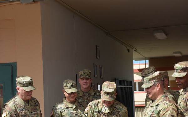 A group of U.S. Army soldiers in OCP (Operational Camouflage Pattern) uniforms are participating in a ribbon-cutting ceremony at a military facility. The ceremony appears to mark the opening or dedication of a building associated with the 94th Army Air and Missile Defense Command, as indicated by unit insignia visible on the columns. One soldier in the center is cutting a red ribbon while others look on smiling.