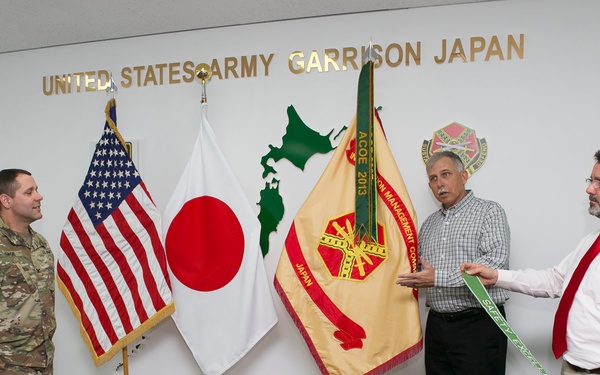 Three individuals are gathered in an official room at United States Army Garrison Japan. Two civilians and one uniformed soldier appear to be participating in a formal presentation or award ceremony involving a green streamer or pennant. The setting includes the US flag, Japanese flag, and a unit guidon flag displayed on the wall behind them.