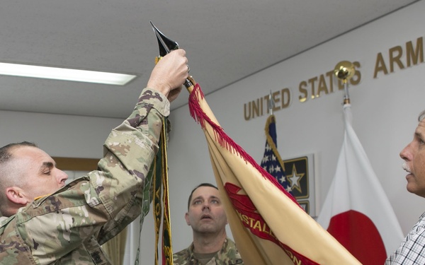 A ceremony is taking place at a United States Army Garrison facility, likely in Japan based on the visible Japanese flag and unit guidon text reading 'JAPAN'. Two soldiers in OCP (Operational Camouflage Pattern) uniforms and a civilian in a plaid shirt are handling a unit guidon/flag, consistent with a change of command or activation/deactivation ceremony. The setting appears to be an indoor conference or ceremonial room.