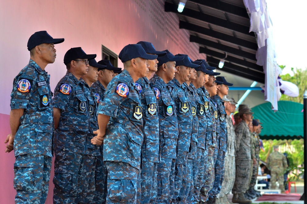 A formation of military or paramilitary personnel dressed in blue digital camouflage uniforms and dark navy caps are standing at attention in a line. They appear to be participating in a formal ceremony or parade, with patches and insignia visible on their uniforms. At the far right, individuals in gray/tan digital camouflage uniforms are also present, suggesting a multinational or joint-service event.