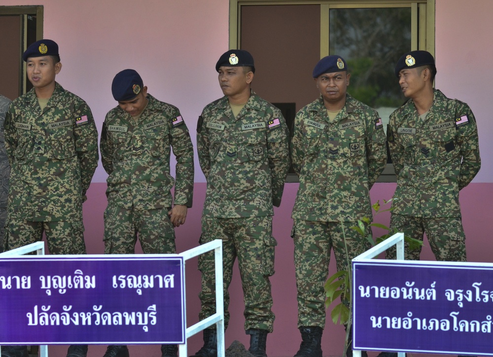 Five Malaysian military personnel are standing in formation in front of a pink building. They are wearing matching digital camouflage uniforms with 'MALAYSIA' name tapes and Malaysian flag patches, along with dark blue berets. Thai-language signage on purple boards is visible in the foreground, suggesting a joint or ceremonial military event in Thailand.