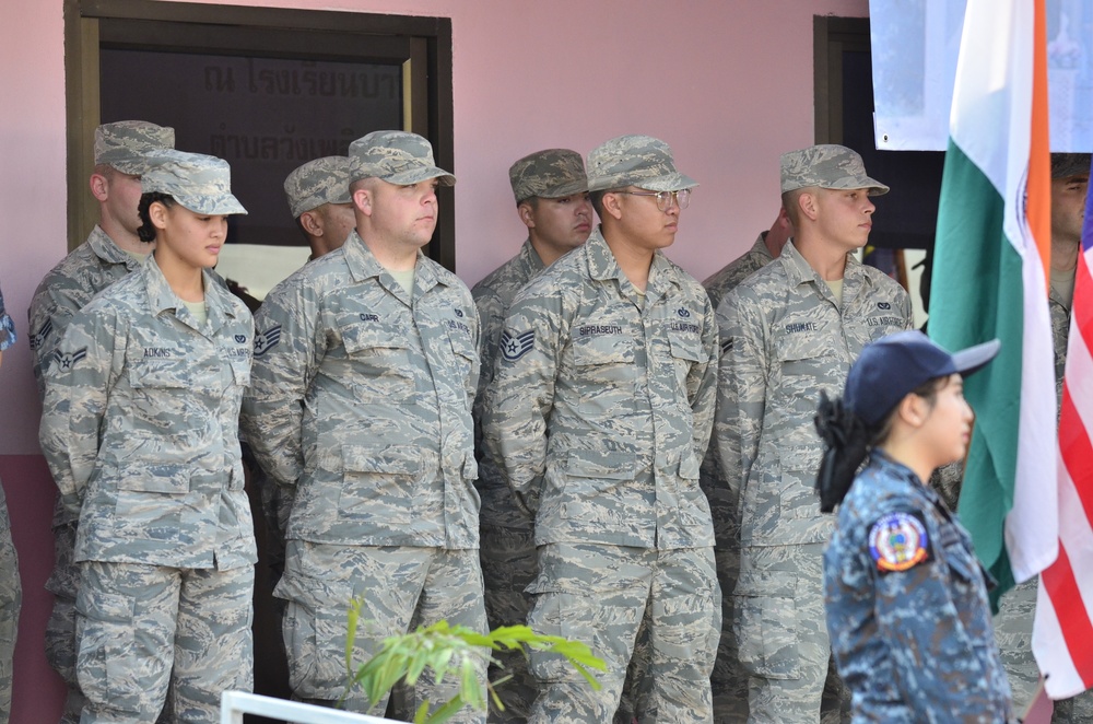 A group of U.S. Air Force personnel in ABU (Airman Battle Uniform) camouflage are standing at attention or at ease during what appears to be a formal military ceremony or formation. Visible name tapes include ADKINS, CARR, SIRPASEUTH, and SHUMATE. A person in a blue uniform, likely from another nation's military, stands in the foreground near flags including what appears to be the Indian flag and the U.S. flag.