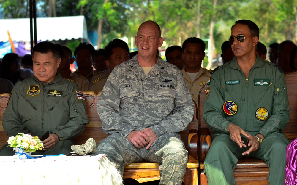 Three military officers from different nations are seated together at what appears to be an official ceremony or event. The central figure wears a U.S. Air Force ABU (Airman Battle Uniform) with the name tape 'FITTS', flanked by officers in olive/sage green flight suits. The setting suggests a multinational military exercise or diplomatic gathering, likely in Southeast Asia.