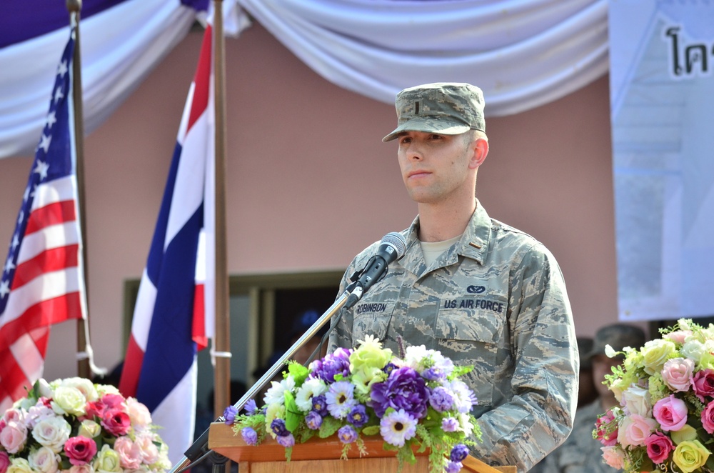 A US Air Force servicemember named Robinson stands at a podium with a microphone, appearing to deliver a speech or address at a formal ceremony. The setting includes both the American flag and what appears to be the Thai flag, suggesting a bilateral military event held in Thailand. Floral arrangements decorate the podium and surroundings, consistent with a ceremonial occasion.