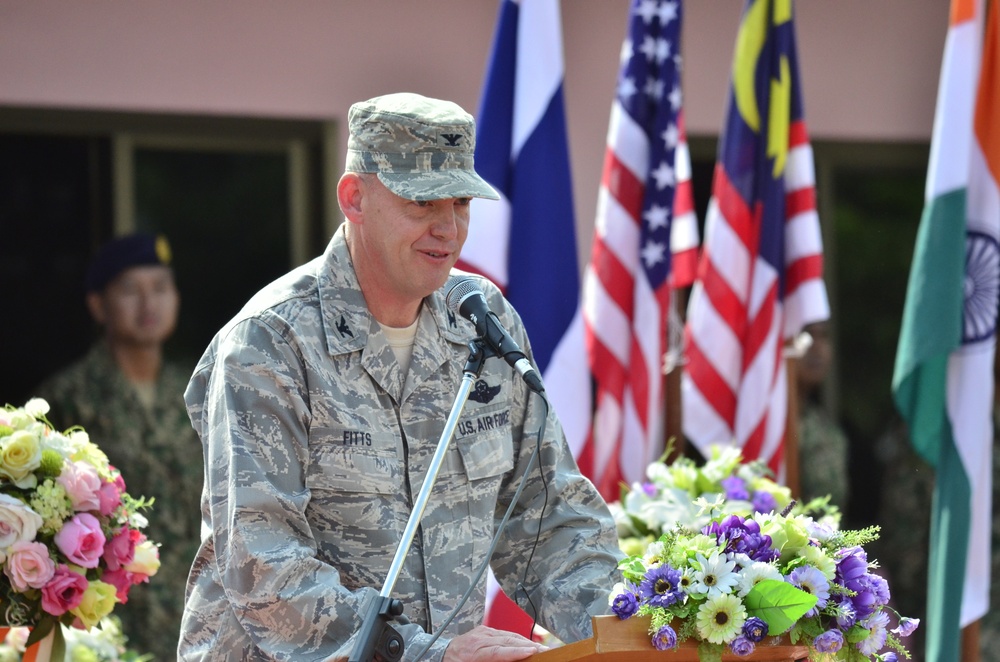A U.S. Air Force officer with the name tag 'FITTS' and colonel rank insignia (eagle) is speaking at a podium with a microphone during what appears to be a multinational military ceremony. Floral arrangements flank the podium, and multiple national flags are visible in the background including those of the United States, Thailand, Malaysia, and India. Additional military personnel in different uniforms are visible in the background.