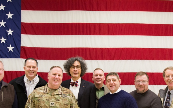A group of nine individuals pose for a formal group photograph in front of a large American flag. One person is wearing U.S. Army camouflage (OCP/MultiCam) uniform with visible rank and name tape, while the others are dressed in civilian attire ranging from business casual to formal. The setting appears to be an indoor military or government facility.