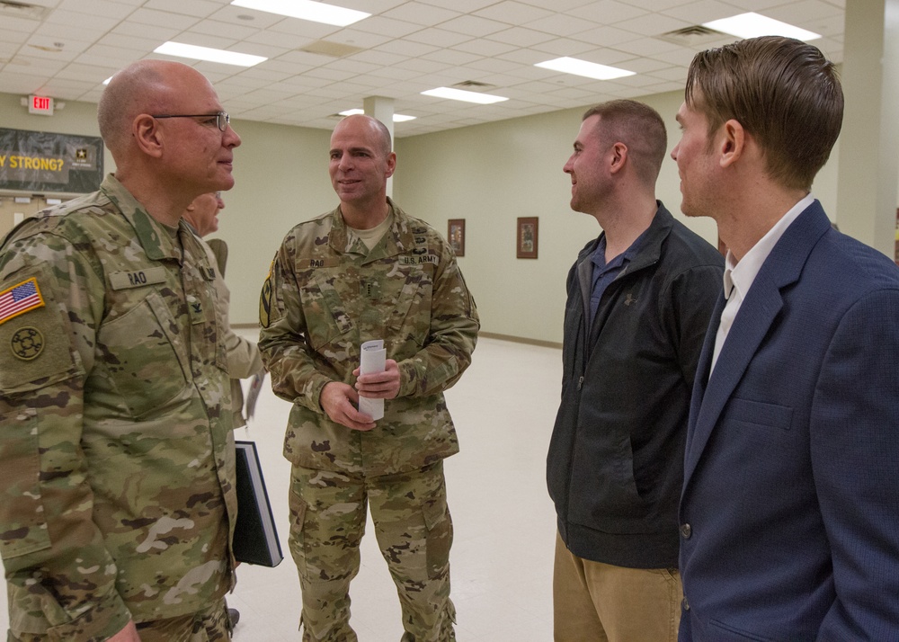 Two U.S. Army soldiers in OCP (Operational Camouflage Pattern) uniforms are engaged in conversation with two civilians in an indoor military facility. The setting appears to be a common area or meeting room within a military installation. The interaction appears informal and conversational in nature.