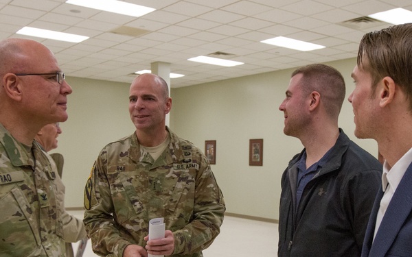 Two U.S. Army soldiers in OCP (Operational Camouflage Pattern) uniforms are engaged in conversation with two civilians in an indoor military facility. The setting appears to be a common area or meeting room within a military installation. The interaction appears informal and conversational in nature.