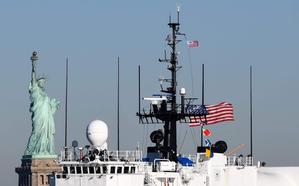 A U.S. Coast Guard cutter with hull number 906 is underway on the water with the Statue of Liberty visible in the background. The large white vessel displays the distinctive Coast Guard diagonal stripe in orange and blue on its bow. The scene appears to be in New York Harbor, with clear skies and calm water conditions.
