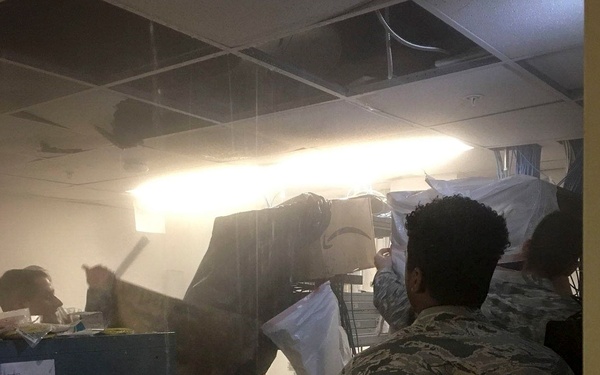 Military personnel in camouflage uniforms (consistent with US Air Force Airman Battle Uniform/ABU pattern) are working inside a damaged room with a partially collapsed drop ceiling. They appear to be covering or protecting rack-mounted equipment with plastic sheeting, suggesting emergency damage control operations. The environment shows signs of water intrusion or structural damage, likely from a storm or similar event.