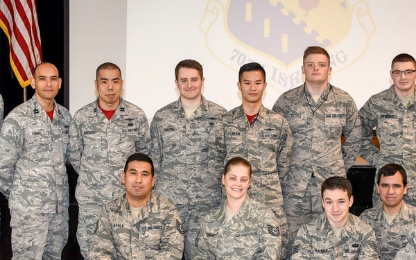 A formal group portrait of approximately 18 United States Air Force personnel in Airman Battle Uniform (ABU) camouflage pattern. The group is posed in two rows — one standing and one kneeling — in what appears to be an auditorium or ceremony hall. An American flag and what appears to be a unit or wing insignia projection screen are visible in the background.