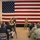 A military briefing or all-hands meeting is taking place indoors before a large American flag backdrop. A uniformed service member appears to be addressing a seated audience of military personnel wearing Navy Working Uniform (NWU) camouflage pattern. A civilian videographer is recording the event with a camera mounted on a tripod.