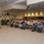 A large gathering of U.S. Navy personnel seated in folding chairs in a hall identified as the 'Hall of Heroes,' likely a military installation auditorium or conference room. Two individuals in Navy camouflage uniforms stand at the front left, apparently addressing the assembled group. The wall behind the audience displays numerous military unit insignia and command seals.