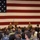 A military briefing or all-hands meeting is taking place in a large hall with a massive American flag displayed on the wall behind the stage. Two uniformed military personnel in camouflage uniforms stand at the front near a wooden podium, addressing a large audience of sailors wearing Navy Working Uniforms (NWU). A video camera on a tripod is positioned to the right, recording the event.