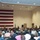 A military ceremony or briefing is taking place in a large indoor hall. A speaker in military uniform addresses a large seated audience composed of military personnel in Navy digital camouflage uniforms and civilians. A large American flag hangs prominently on the left wall, and a military unit flag is visible near the podium.
