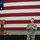 A U.S. Navy officer in digital camouflage (NWU Type III) uniform stands on a stage speaking before a large American flag backdrop. He is positioned to the right of a wooden podium equipped with a microphone. The setting appears to be a formal military all-hands or briefing event held in a large indoor facility such as a hangar or gymnasium.