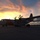 A US Coast Guard HC-130 Hercules aircraft (tail number 2007) is parked on a tarmac at sunset or sunrise, silhouetted against a dramatic orange and blue sky. The aircraft displays the distinctive Coast Guard white and orange livery. A small group of personnel are gathered near the aircraft's fuselage, likely conducting pre-flight or post-flight activities.