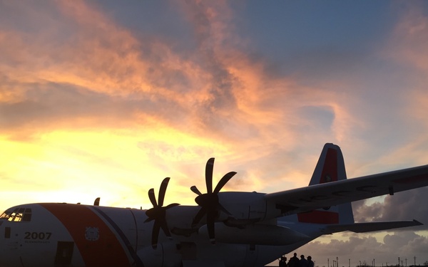 A US Coast Guard HC-130 Hercules aircraft (tail number 2007) is parked on a tarmac at sunset or sunrise, silhouetted against a dramatic orange and blue sky. The aircraft displays the distinctive Coast Guard white and orange livery. A small group of personnel are gathered near the aircraft's fuselage, likely conducting pre-flight or post-flight activities.